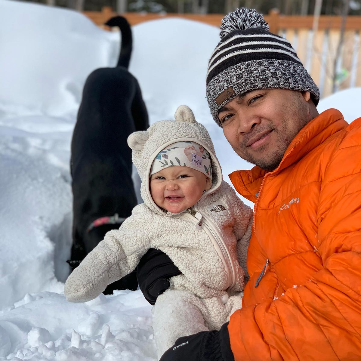 Photo of Jun Robinson holding a baby wearing an animal-themed onesie and knelt in the snow, a black dog with a red collar in the background with its nose in the snow.