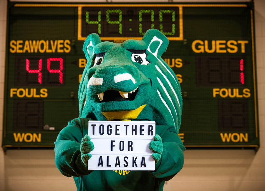UAA mascot holds a Together For Alaska white sign in front of a scoreboard