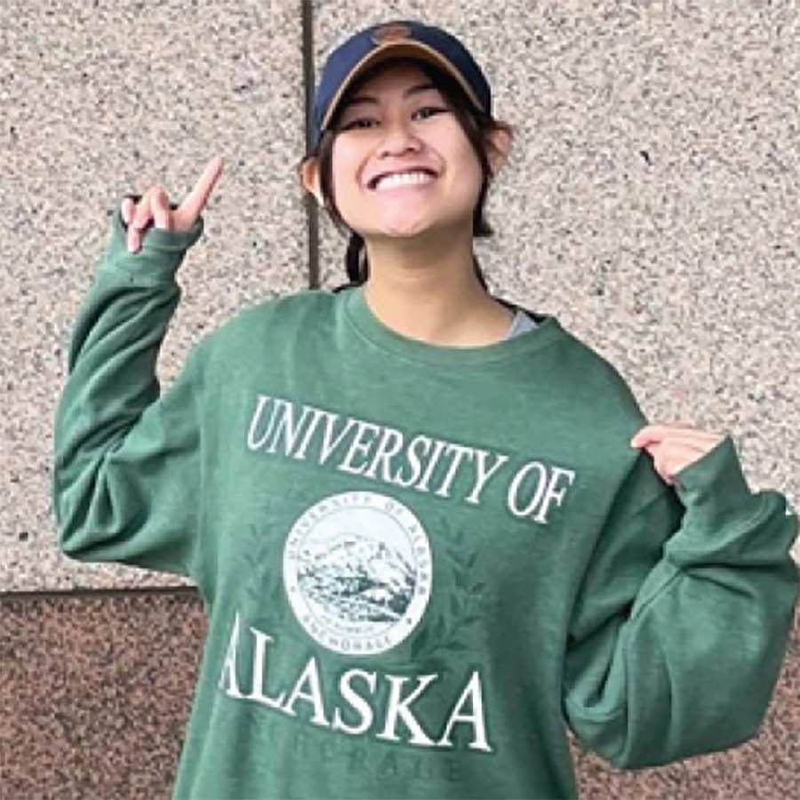 A portrait photograph of Christina-May Castro smiling in a navy blue hat and green University of Alaska sweatshirt posing for a picture with her right index finger pointed upward
