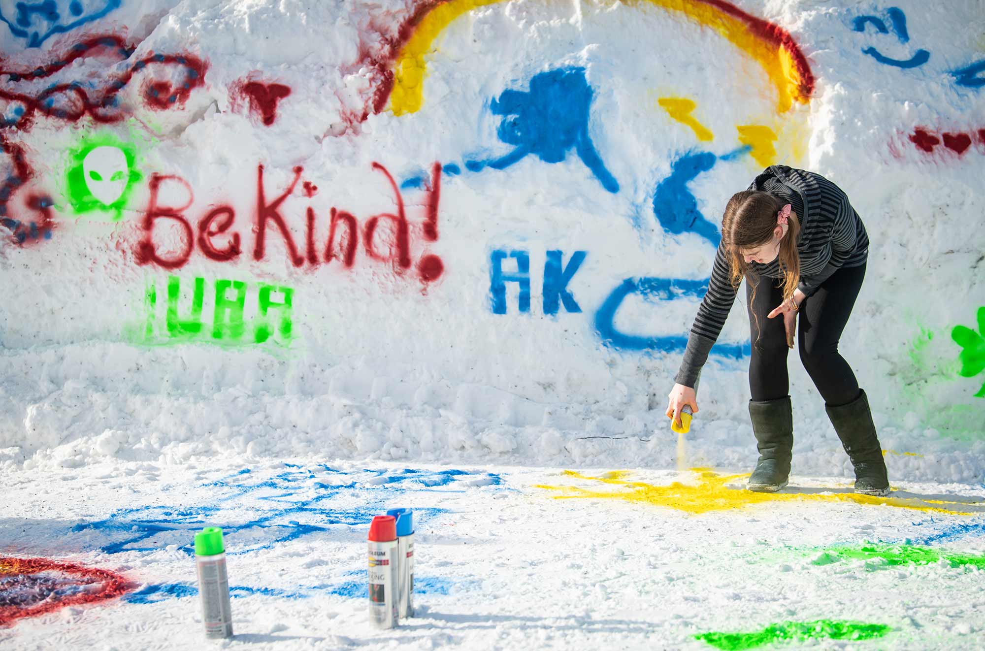 A woman in a dark gray and black striped jacket plus black sweatpant leggings with dark olive green boots sprays down a yellow dye color onto some snow on the ground with her finger pressed onto the top of a chrome colored aluminum canister bottle as she decorates all over the snow many different types of graffiti artwork symbols such as a green backdrop alien, phrases such as "Be Kind!", "UAA", "AK", and other variety of shapes (state of Alaska), etc. while there are other chrome colored aluminum canister bottles nearby in green, red, and blue dye colors as part of UAA’s Winterfest Art & Music event in the Cuddy Quad in February 2023.