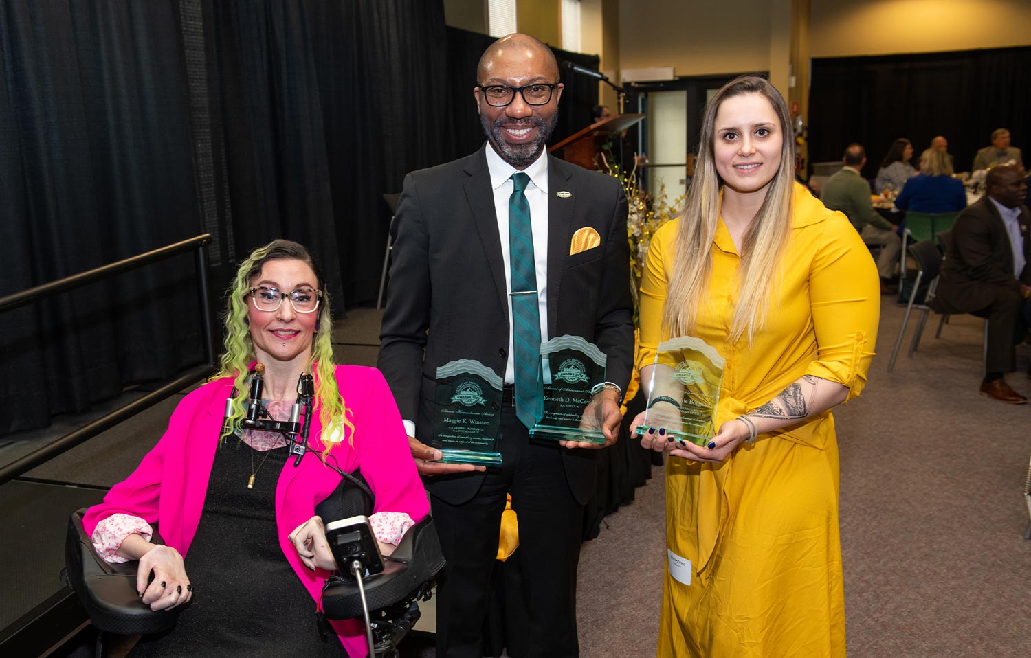 Maggie Winston wearing a black dress and a hot pink blazer (left), Kenneth McCoy wearing a black suit and blue tie(center) and Marie-Sophie Boggasch wearing a vibrant yellow dress (right) stand near the stage holding their glass awards at the 2022 Alumni of Distinction Celebration Banquet 