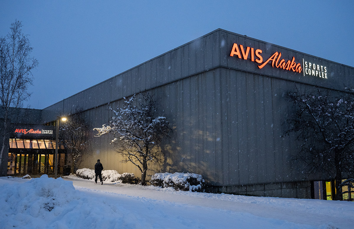Person walking into the Avis Alaska Sports Complex building in the evening, snow covering the ground