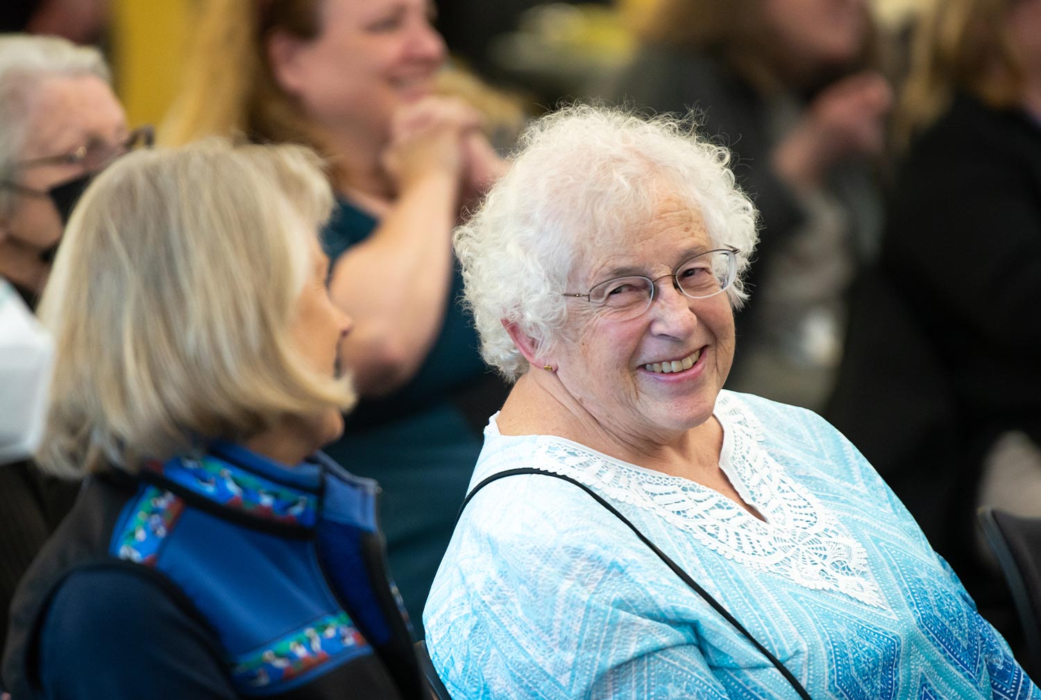 seated in an audience, Nancy Lesh smiles while looking at a woman seated to her right