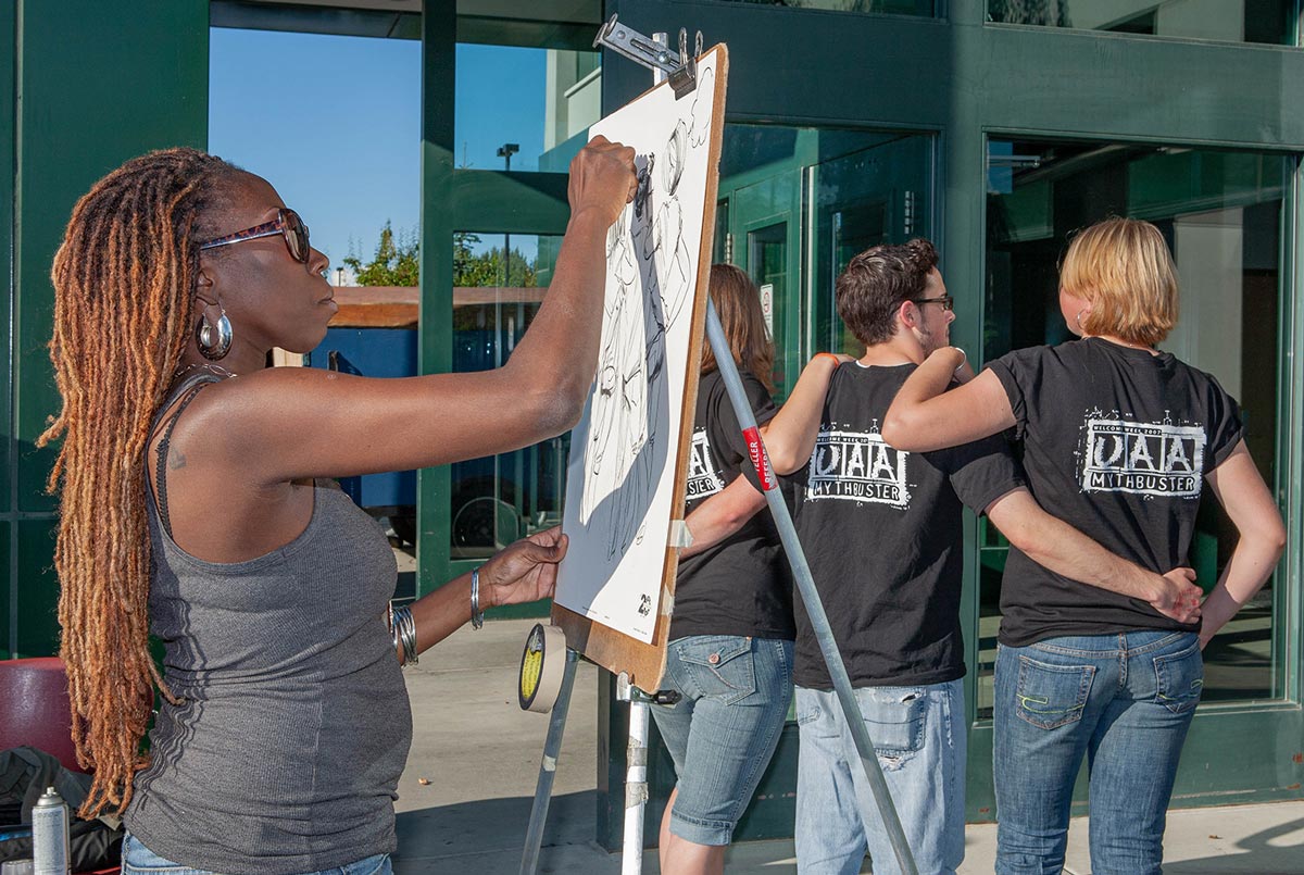 a black woman with long orange-red locs stands, sketching three subjects wearing matching UAA shirts and facing away from the artist