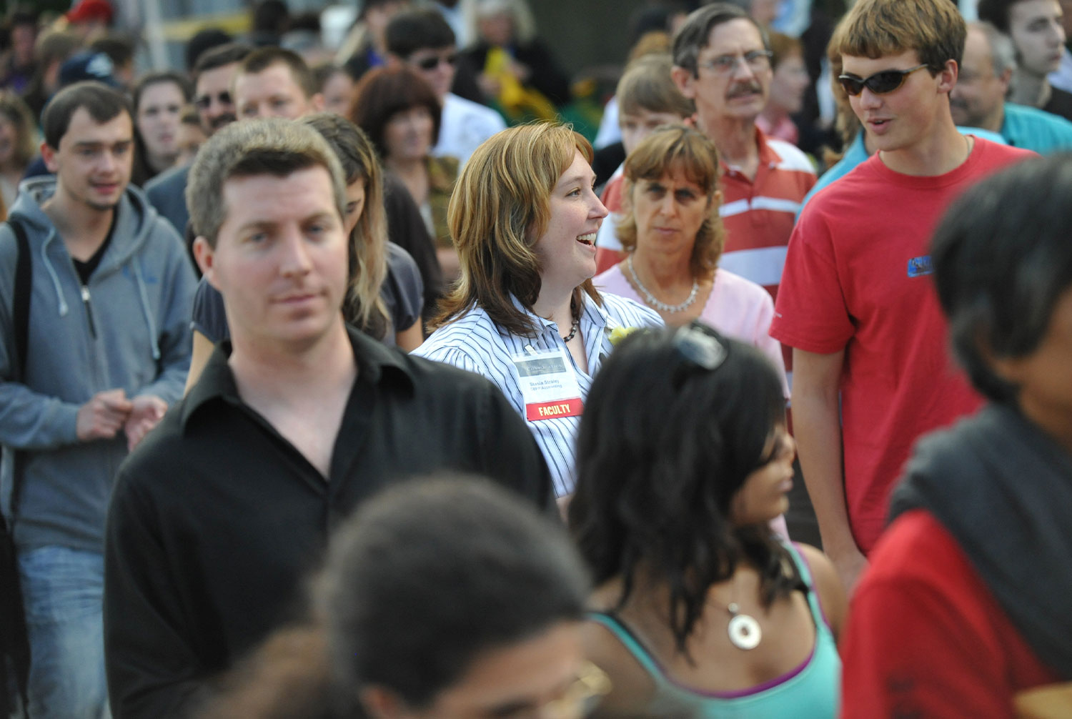 a woman wearing a faculty badge clipped to her collar talks to a young man while standing in a large crowd