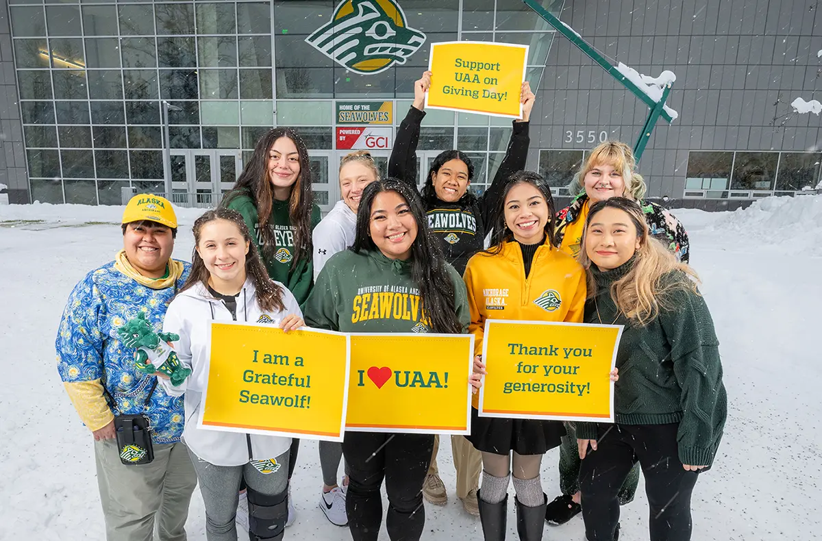 Landscape close-up photo outdoor view of UAA supporters standing in snow-covered ground at the Alaska Airlines Center south entrance area as they are smiling in their respective UAA attire holding the school's gold color Giving Day small poster signs with different messages in the school's green color that say Support UAA on Giving Day, I am a Grateful Seawolf!, I love UAA!, and Thank you for your generosity!