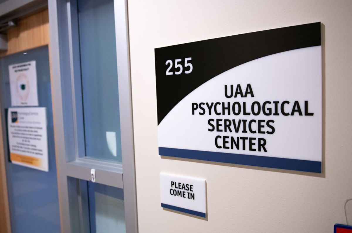 Landscape close-up photo indoor view of the UAA Psychological Services Center entrance sign displayed on the wall with another smaller sign underneath that which reads Please Come In (both of these signs are pictured to the far right) and next to those signs pictured to the far left is a closed entrance door with two placard laminated for your information detail signs on it
