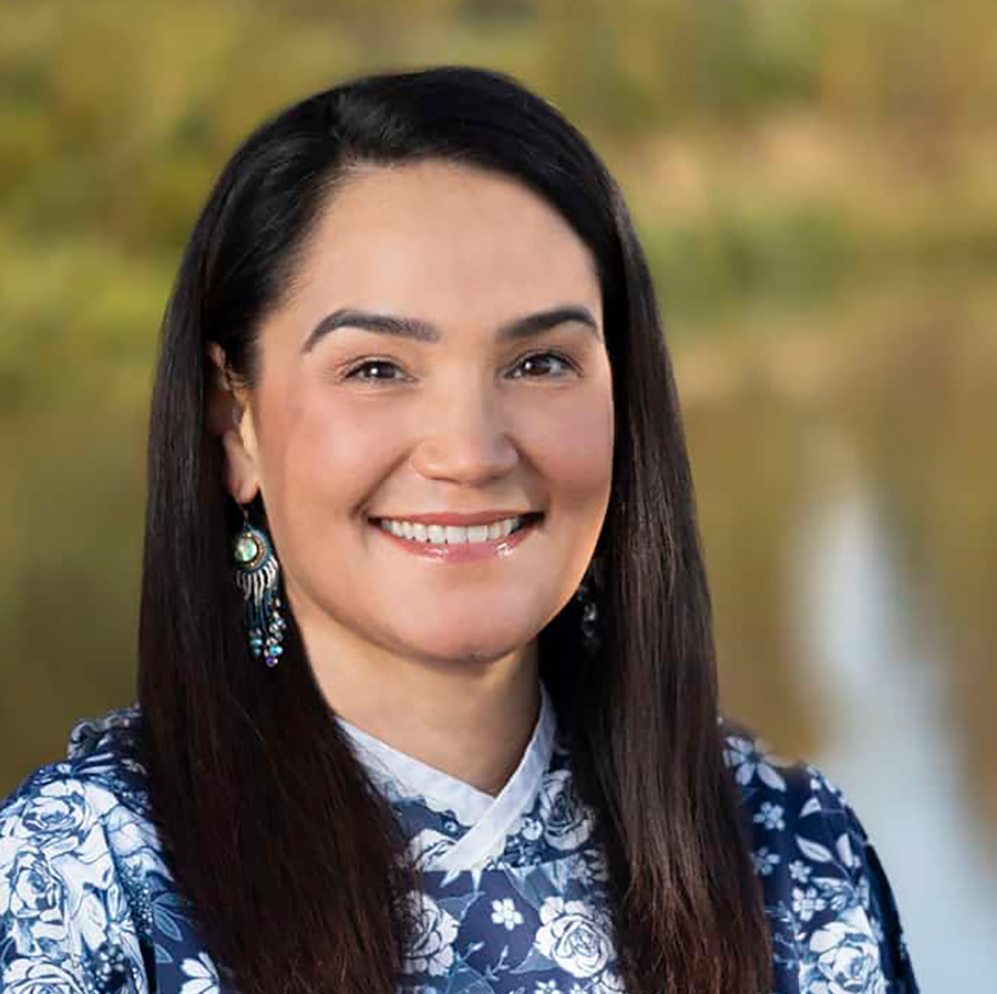 Portrait close-up photo view of Sarah Lukin smiling in a custom-made white/blue floral rose pattern themed dress and wearing tribal shaped themed earrings