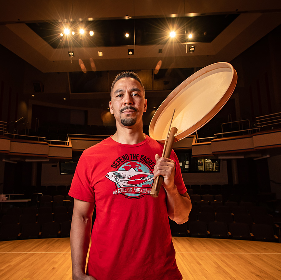 Portrait close-up photo view of Phillip Blanchett grinning in a red graphic t-shirt that has a whale and earth shaped illustration theme on it that has a message saying Defend The Sacred: Our Bodies, Our Lands, Our Ways as he is holding a large, round ceremonial drum and a drumstick and he is standing on a performing arts theater stage indoors with bright stage lights around and empty rows of seats in the far distance