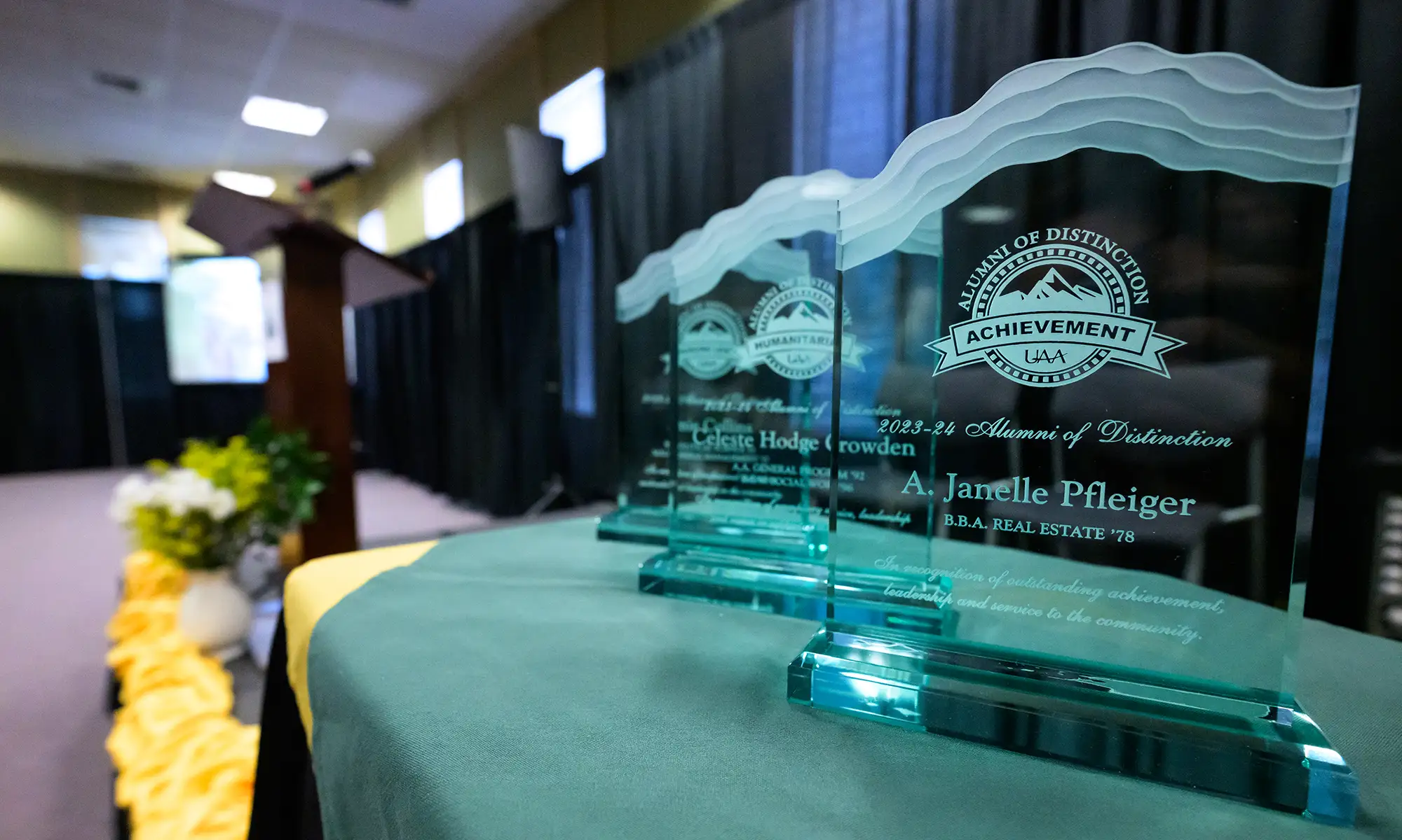 close view of three glass UAA Alumni of Achievement trophies awarded to Amie Collins, Celeste Hodge Growden, and A. Janelle Pfleiger lined on a table; a podium and small stage are visible in the background