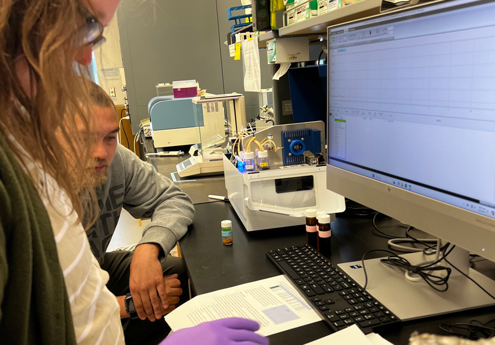 Two individuals examine data on a computer monitor in a lab, surrounded by scientific equipment and documents.