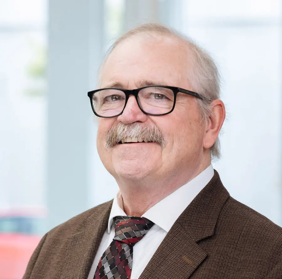 Portrait headshot close-up photo view of Joseph Levesque smiling in outer frame black trim see through prescription glasses and a dark brown plaid business suit blazer and white button-up dress shirt underneath with a multi-colored (red, black, dark grey diamond shape style pattern) tie