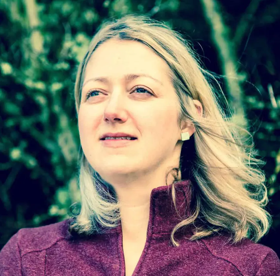 Portrait headshot close-up photo view of Kelly Harrell grinning in a dark magenta jacket with chrome square shaped earrings