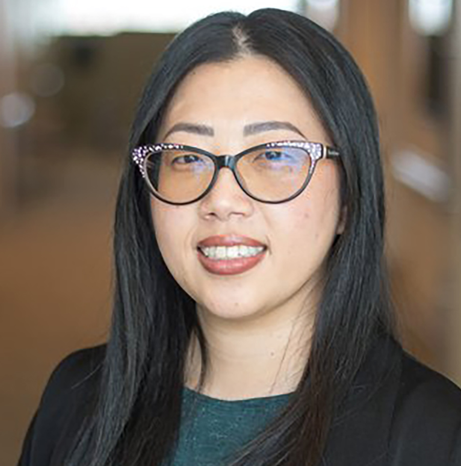 Portrait orientation close-up headshot photograph of By Thao smiling in a black business blazer suit with a dark forest green t-shirt underneath and dark tinted outer frame see through prescription see-through eyeglasses