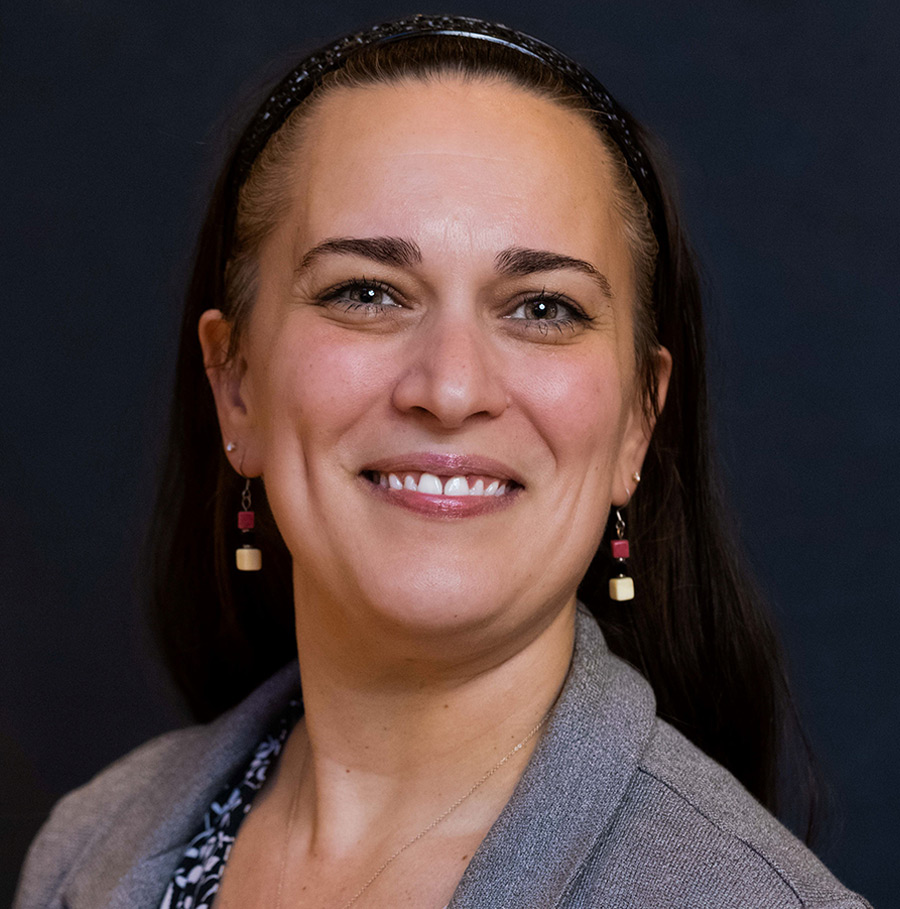 Portrait orientation close-up headshot photograph of Christine Hopkins smiling in a dark grey business suit blazer with a multi-colored (black and white) blouse underneath as she has on square shaped earrings that contain multiple different colors (red and creamy off-white tan)