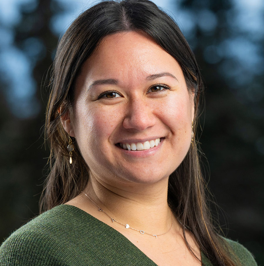 Portrait orientation close-up headshot photograph of Pearl-Grace Pantaleone smiling in a dark forest green cardigan sweatshirt with a thin chrome necklace equipped around her and custom-shaped design earrings