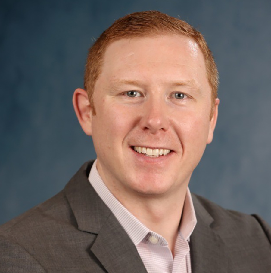 Portrait orientation close-up headshot photograph of Robert Champion smiling in a dark grey business blazer suit and a light faded pink button-up dress shirt underneath