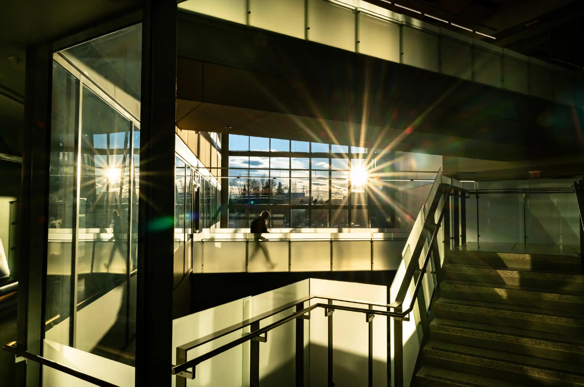 Portrait orientation interior photograph view of a low winter sun shining into the atrium of UAA’s ConocoPhillips Integrated Science Building through large windows, casting strong shadows on the walls as there visibly happens to be a person walking along a walkway path