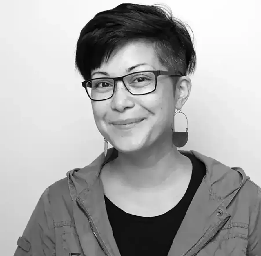 Headshot black and white photograph of Asia Buzon grinning in a t-shirt and jacket with prescription eyeglasses equipped as well as earrings