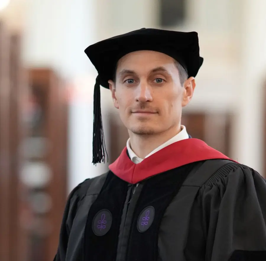 Headshot photograph of Corey Hester grinning in a black graduation gown outfit with a cap as well as a red ribbon accessory around his neck