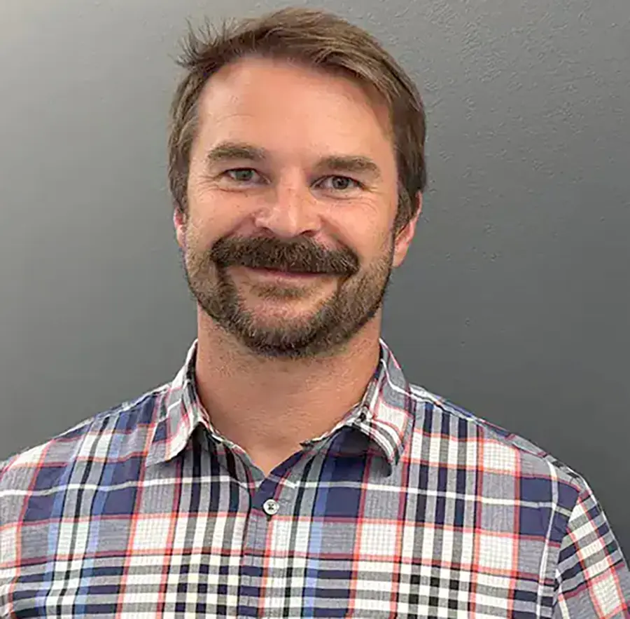 Headshot photograph of Jason Johnston grinning in a blue/white/black/red button-up dress shirt