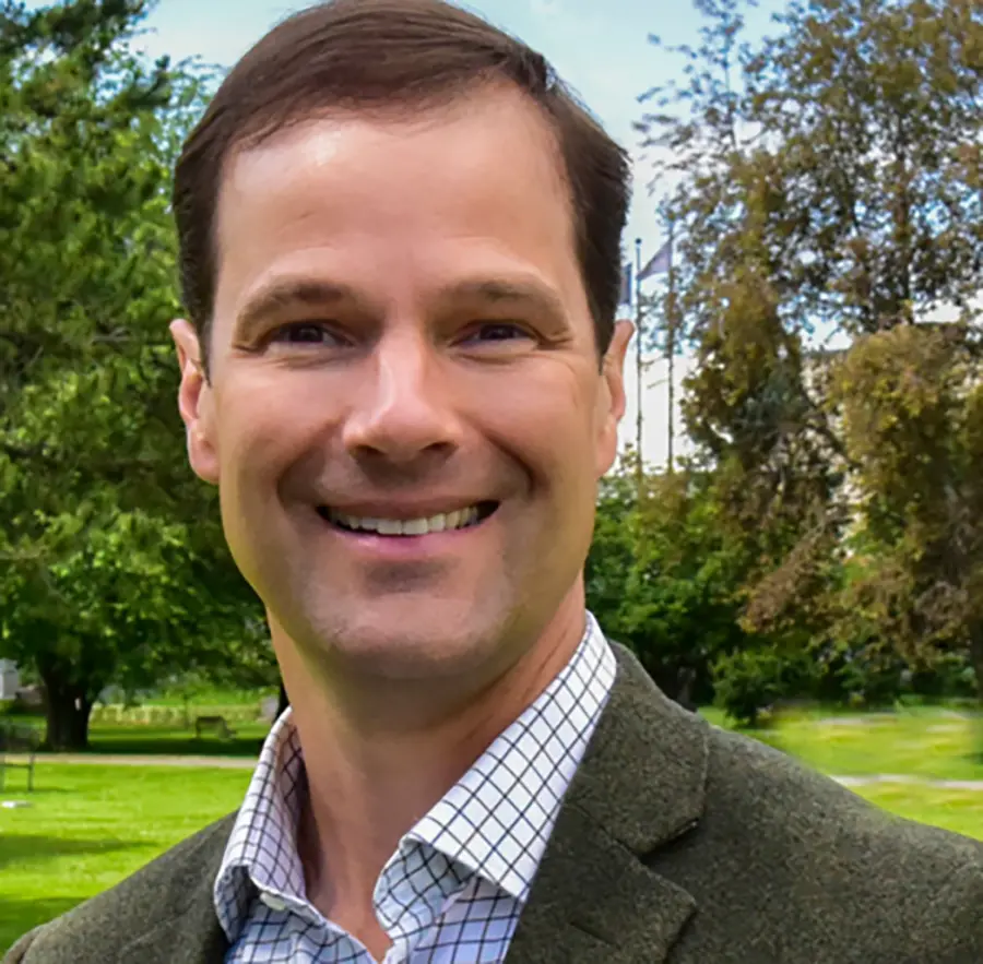 Headshot photograph of Kevin Pendergast smiling in a dark olive green blazer business suit with a white/black vertical linear pattern style button-up dress shirt underneath