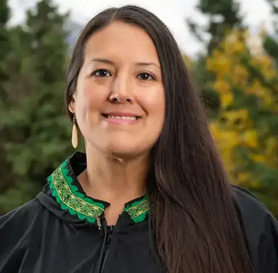 Headshot photograph of Michele Yatchmeneff smiling in a black jacket with a green/yellow cultural tribal style design collar accessory around nearby her neck area