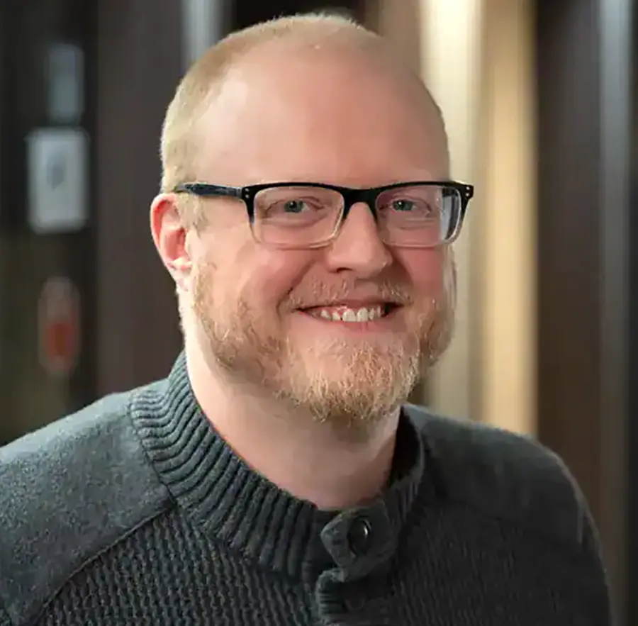 Headshot photograph of Toben Shelby smiling in a dark grey button-up cardigan turtleneck sweatshirt with prescription eyeglasses equipped