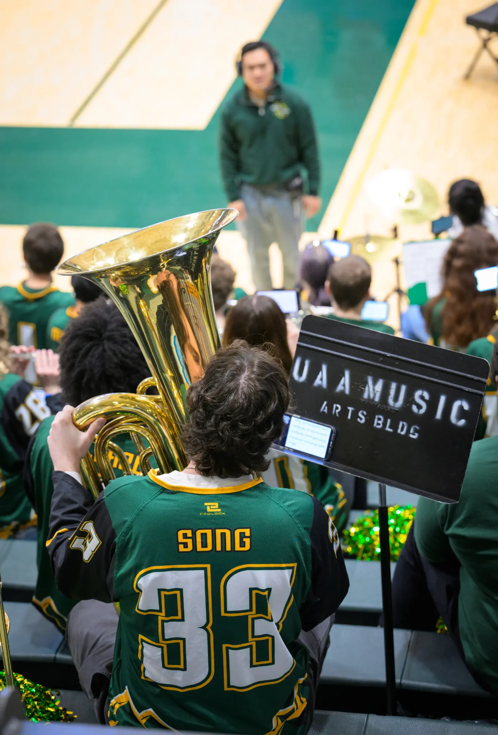 A tuba player, who happens to be a male UAA (University of Alaska Anchorage) person, is seen from the back seated on the bleachers with other members of the UAA Pep Band or orchestra inside UAA's Alaska Airlines Center; The tuba player is wearing a green and black jersey that says the word or last name SONG and the numeral 33; A black sign music stand nearby reads UAA MUSIC ARTS BLDG; A conductor is visible, out of focus, in the background