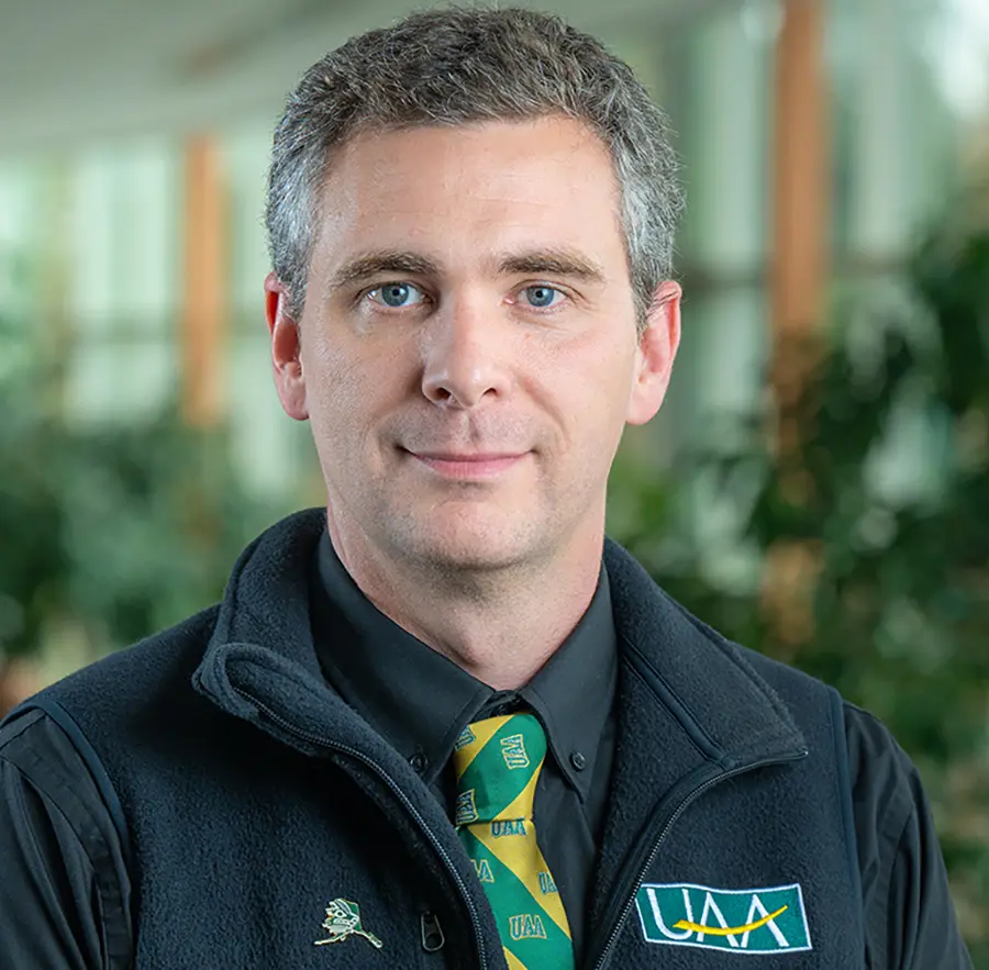 Headshot photograph of Zac Clark grinning in a University of Alaska Anchorage (UAA) black vest/jacket with a black button-up dress shirt underneath alongside a UAA school green/yellow colored tie