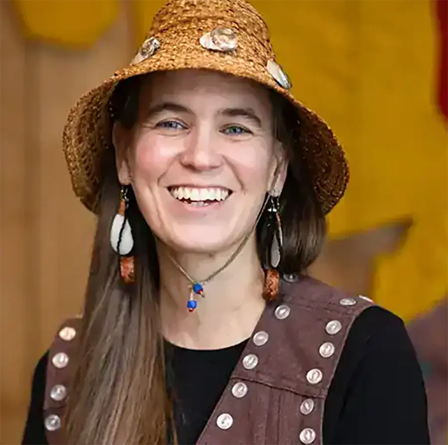 Headshot photograph of Karla Gatgyedm Hana’ax Booth smiling in a black sweatshirt with a brown vest and a woven straw hat
