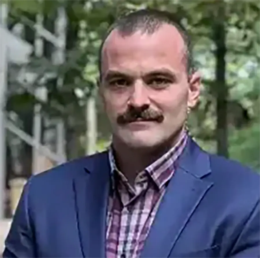 Headshot photograph of Kris Squires grinning in a navy blue business blazer suit