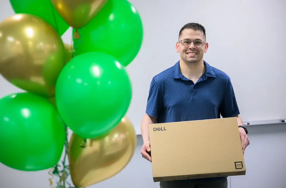 David Murray smiles in a navy blue polo top holding a new Dell laptop product box with green and gold color balloons nearby