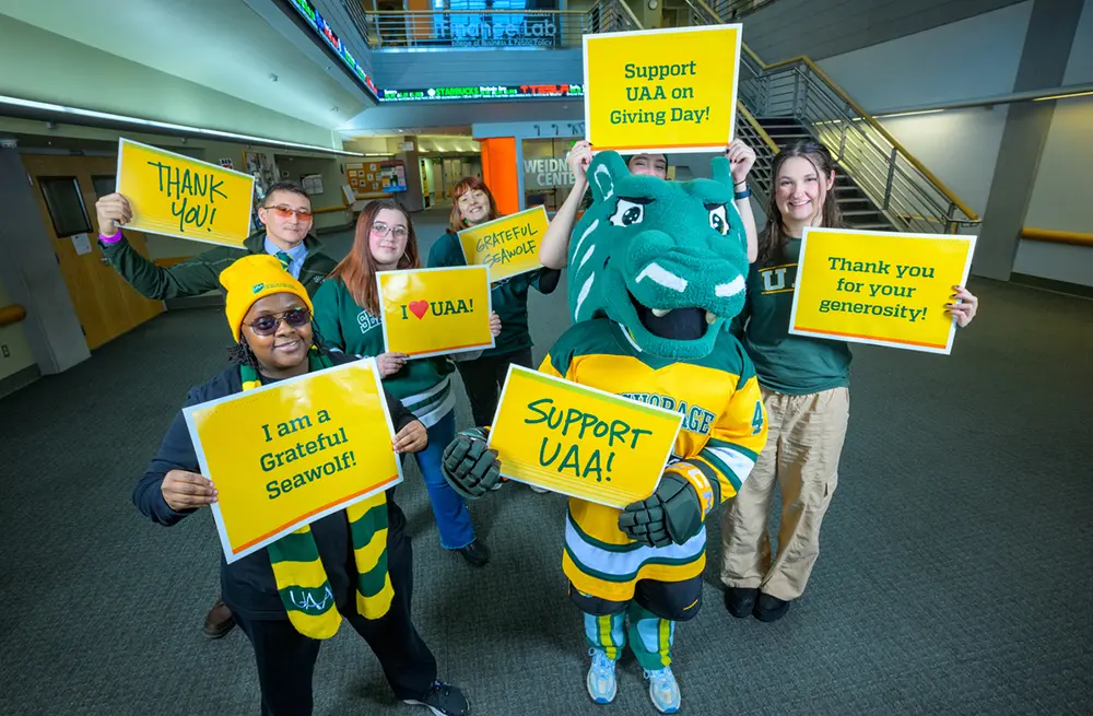 Group of six students and the UAA Spirit the Seawolf mascot stand in a campus lobby holding yellow and white signs for UAA Giving Day