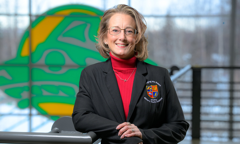 Deborah Morton, wearing a red turtleneck and a black blazer with a crest, smiles and leans against a railing. A large green and yellow Seawolf logo is visible in the background.
