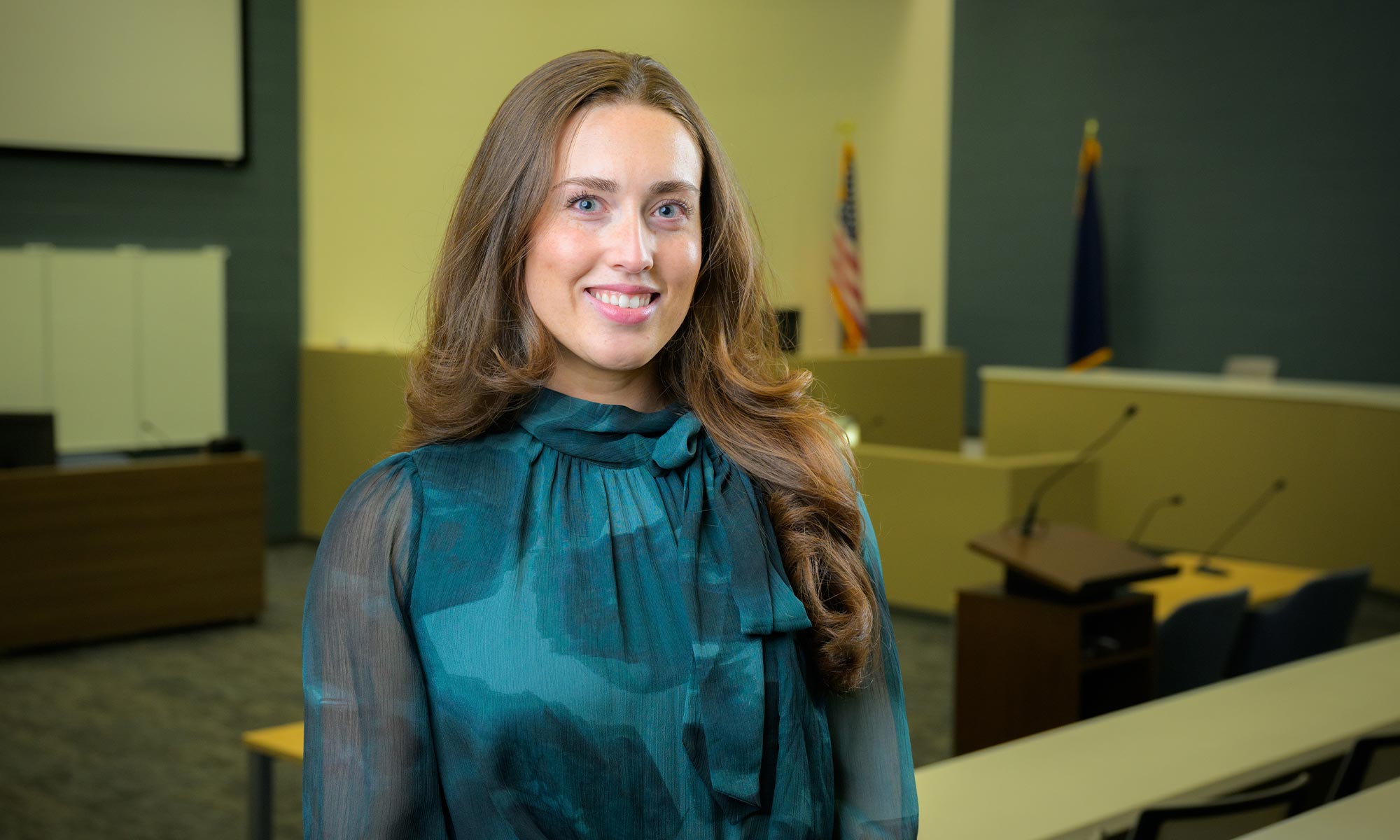 Maile Johnston, wearing a teal patterned blouse with a high neckline, smiles for a portrait. She stands in a room with long tables and podiums that resembles a lecture hall or courtroom.