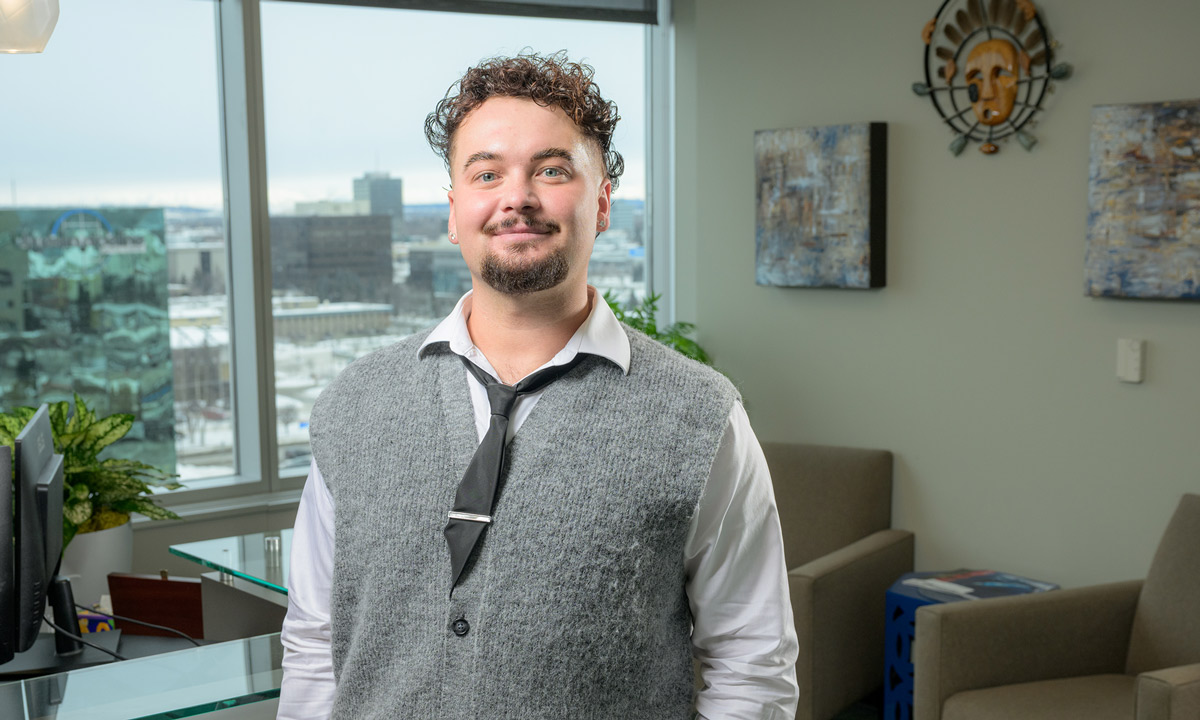 Vladislav Finko, wearing a grey sweater vest over a white collared shirt and black tie, smiles in an office setting. A large window behind him shows a view of a city skyline under a grey sky.